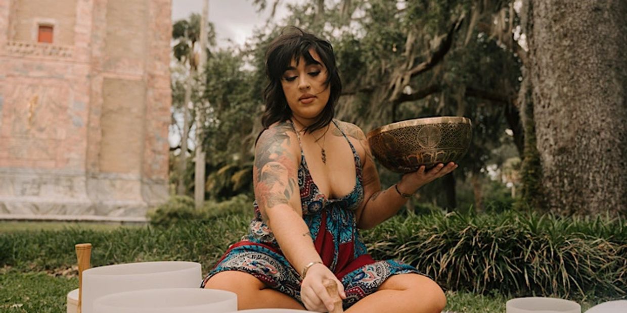 Woman with tattoos plays crystal singing bowls outdoors in a peaceful setting.