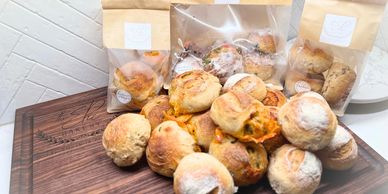 Assorted freshly baked bread rolls on a wooden board with packaged sets in the background.