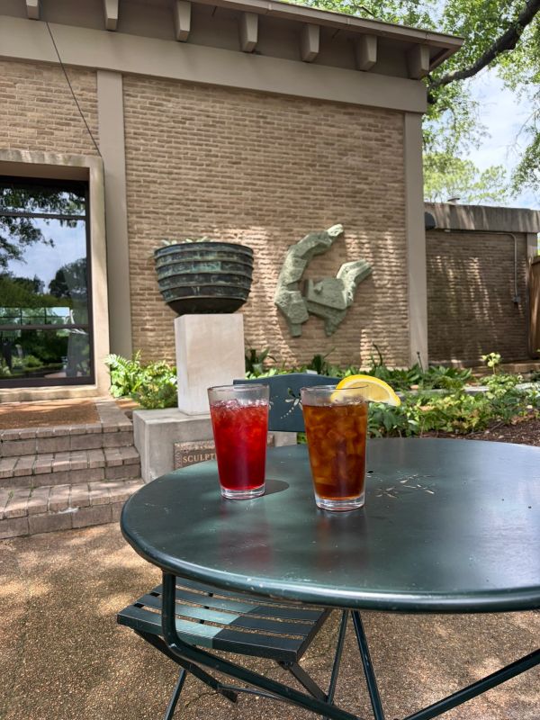 Two iced drinks on a round outdoor table with a brick wall and sculpture in the background.