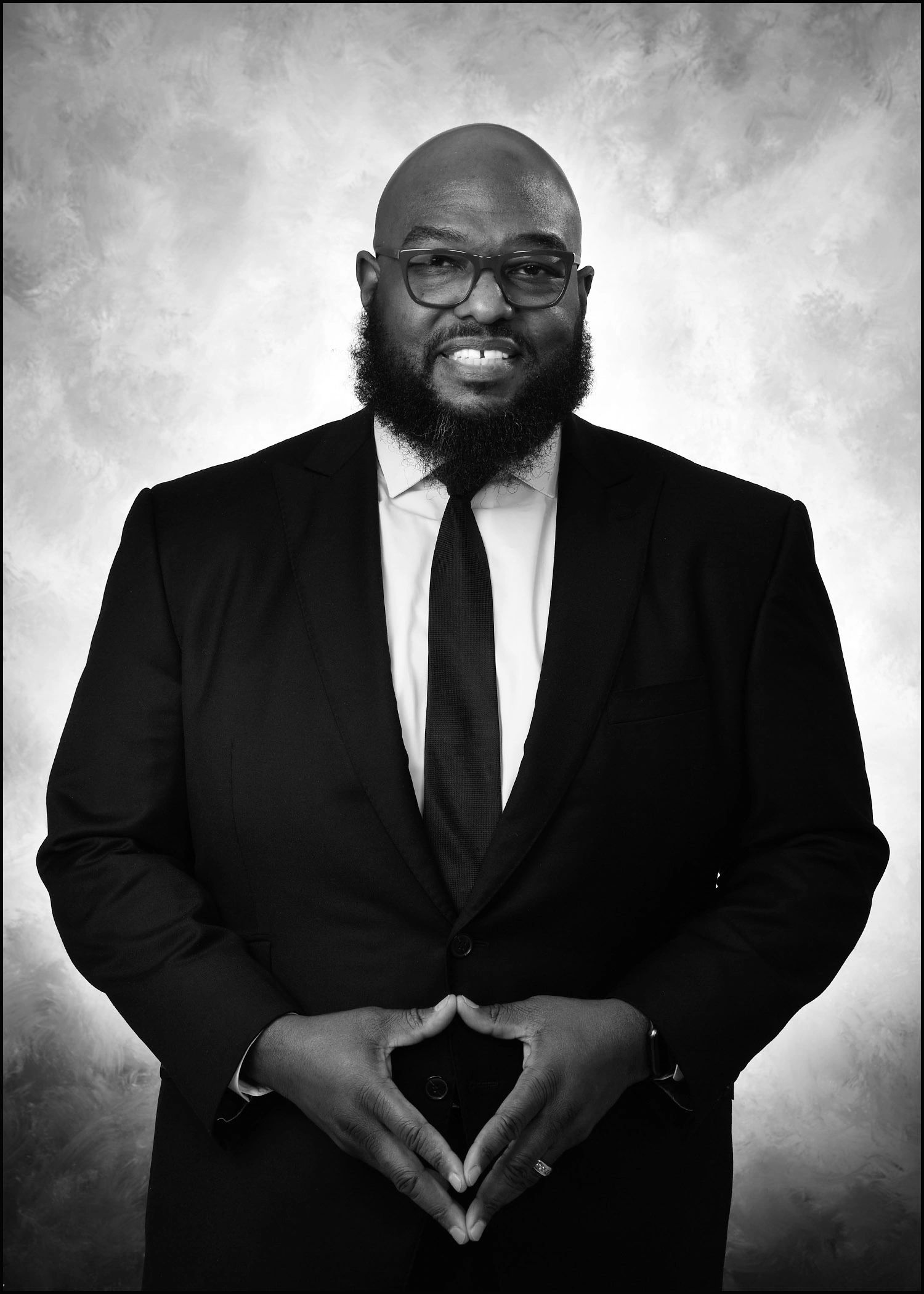 Smiling man in a black suit and tie with glasses.