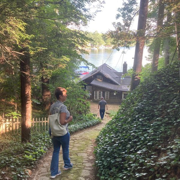 Two people walking down a forest path toward a lakeside cabin.
