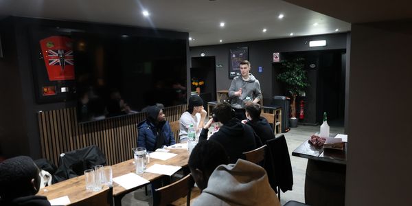 A group meeting in a modern room with a speaker addressing attendees.