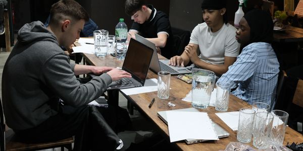 Four young men working together at a table with laptops and notes.