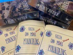 Stacks of Holy Bibles with handwritten covers saying 'Faith Can Move Mountains.'