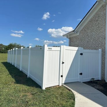 A long white vinyl fence encloses a backyard beside a brick house under a blue sky.