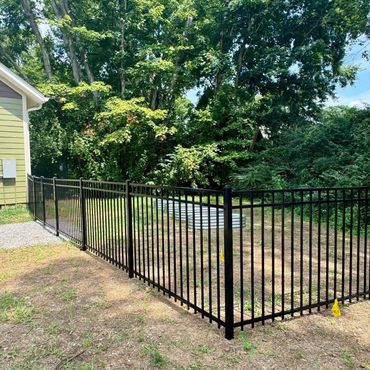 Black metal fence enclosing a yard beside a green house and trees.