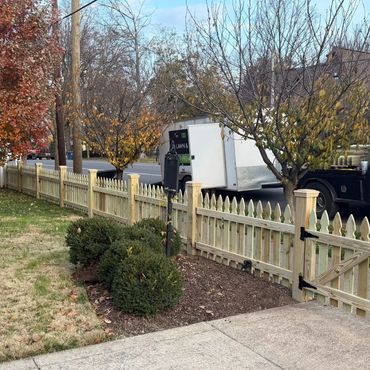 New wooden picket fence with autumn trees and landscaping in a residential neighborhood.