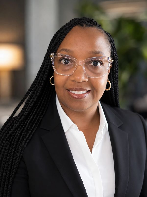 Professional woman with braided hair and glasses wearing a black blazer and white shirt.