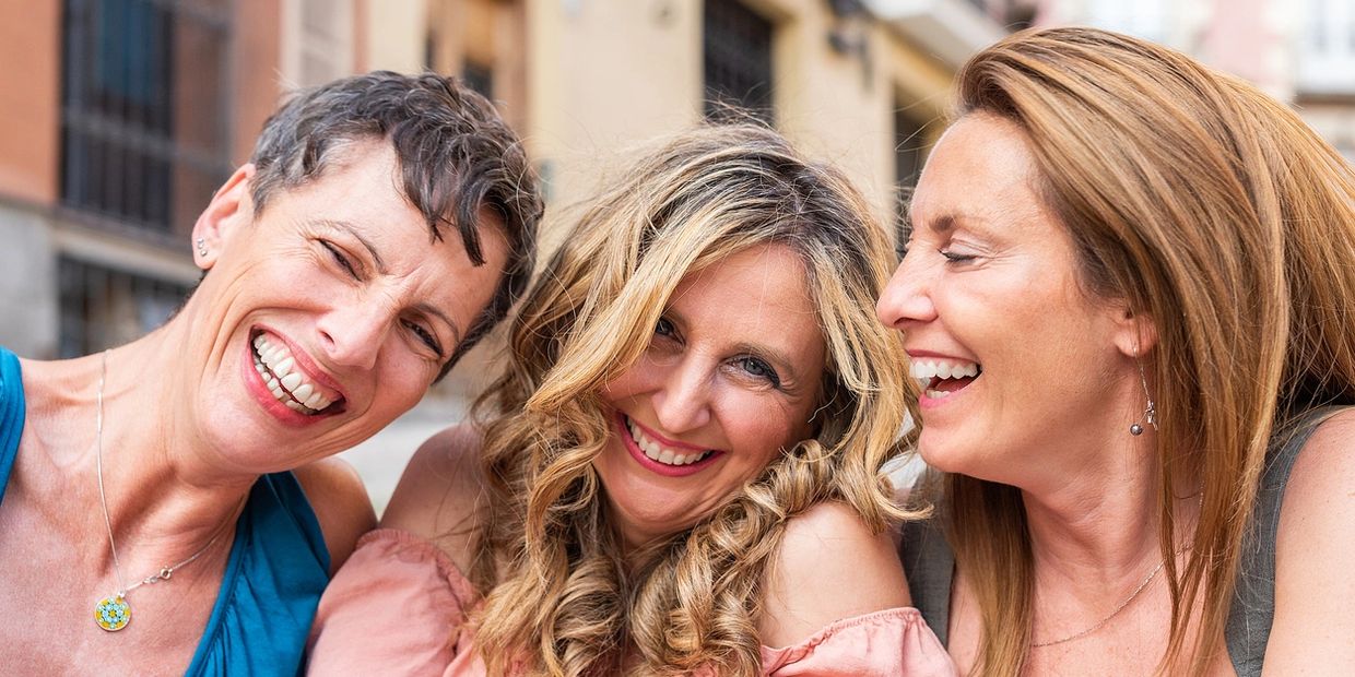 Three women smiling and enjoying a joyful moment together outdoors.