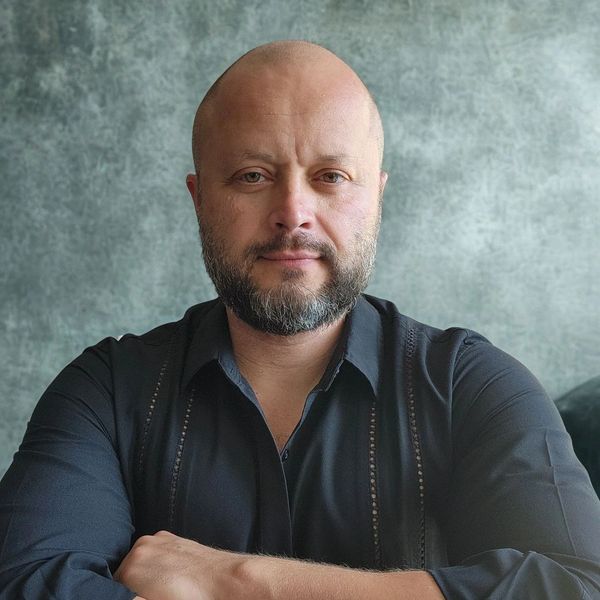 A confident bald man with a beard wearing a black shirt against a textured gray background.