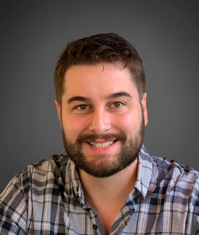 Smiling man with beard wearing a plaid shirt against a gray background.