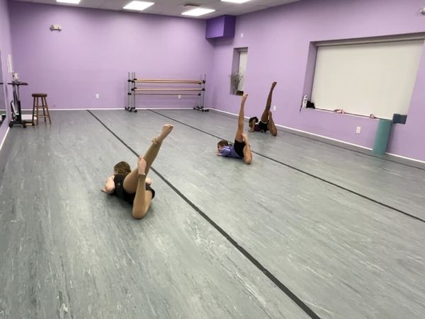 Two dancers practicing leg lifts in a purple-walled studio.