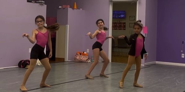 Three young girls dancing in a studio wearing pink tops and black bottoms.