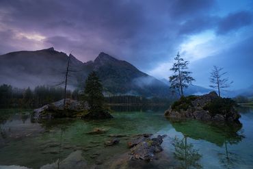 Ein atmosphärisches Landschaftsfoto des Hintersees in den Berchtesgadener Alpen bei Morgendämmerung. Im Vordergrund ist das kristallklare, türkisgrüne Wasser des Sees zu sehen, aus dem charakteristische Felsinseln mit einzelnen Nadelbäumen herausragen. Die Felsen sind mit Moos bewachsen und spiegeln sich im ruhigen Wasser. Im Hintergrund erheben sich die majestätischen Gipfel der Alpen, teilweise in Morgennebel gehüllt. Der Himmel zeigt ein dramatisches Farbenspiel aus violetten und blauen Tönen mit ziehenden Wolken. Die herbstliche Stimmung wird durch die kargen Bäume auf den Felsinseln unterstrichen. Die Szene strahlt eine mystische, friedvolle Ruhe aus, typisch für die frühen Morgenstunden in den Bergen.