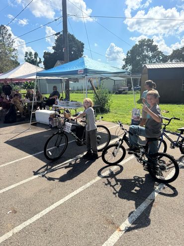 Two boys on bikes at an outdoor event with tents.