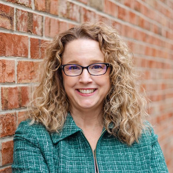 Smiling woman in professional attire with curly hair and glasses standing against a brick wall.