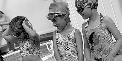 Three young girls in swimwear and swim caps preparing for a swim.