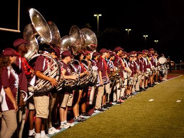 High school marching band in maroon uniforms lined up on football field at night.