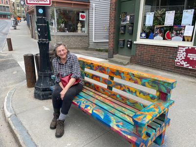 Deborah sitting on a colorful bench painted by her son, Peter Bird, located on Broadway in Saranac L