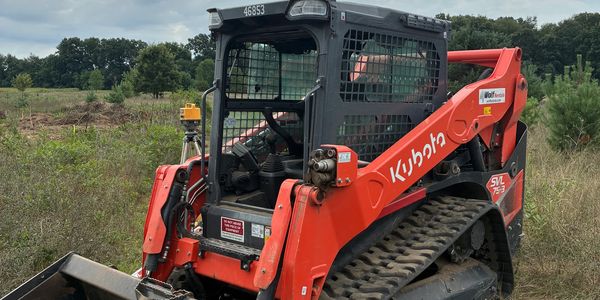 An orange Kubota SVL75-3 compact track loader on a grassy field under a cloudy sky.