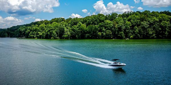 A motorboat speeds across a lake surrounded by lush green trees under a blue sky.