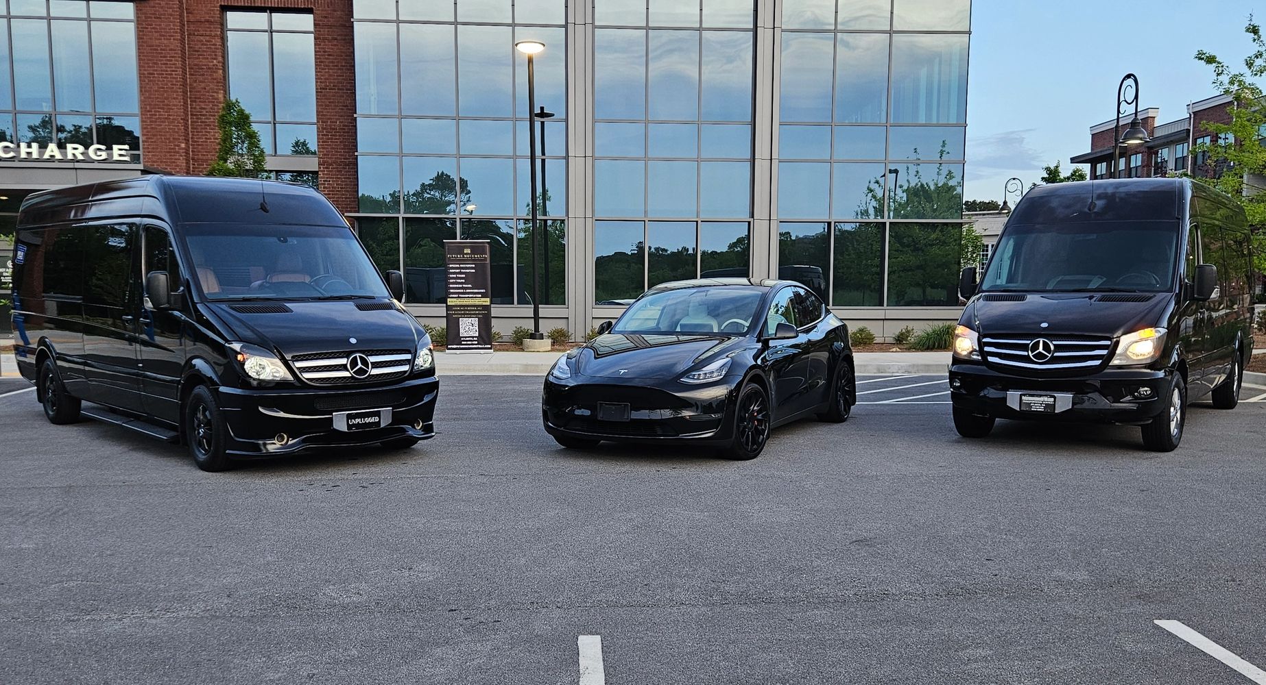 Three black vehicles, two Mercedes vans and one Tesla car, parked in front of a glass building.