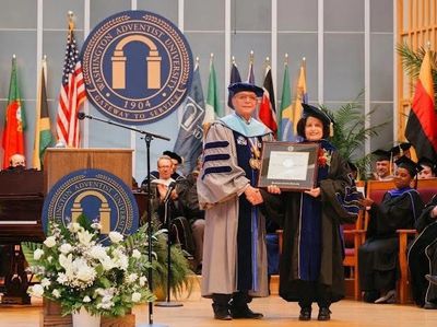 Two graduates in academic regalia holding a diploma on stage.