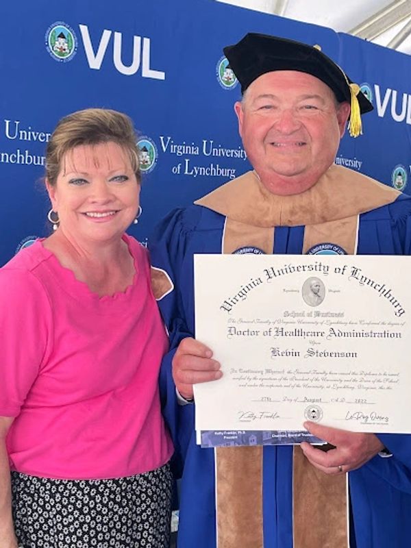 Man in graduation gown holding diploma, woman standing next to him.
