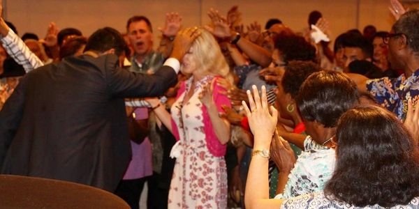 A man places his hand on a woman's head during a spiritual gathering with many people praying.