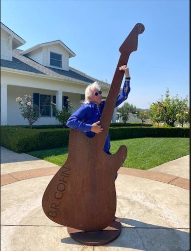 Person posing with a giant guitar sculpture outside a house.