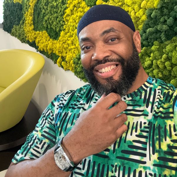 Smiling man with a beard wearing a patterned shirt and watch, seated by a green and yellow moss wall.