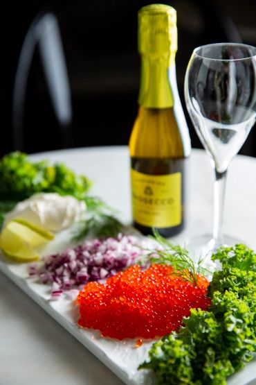 Red caviar with greens, lemon, onion, prosecco bottle, and empty glass.