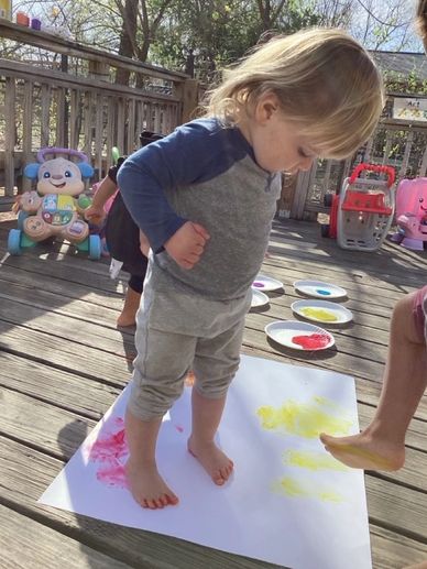 A toddler stands on paper painting with feet outdoors on a sunny deck.