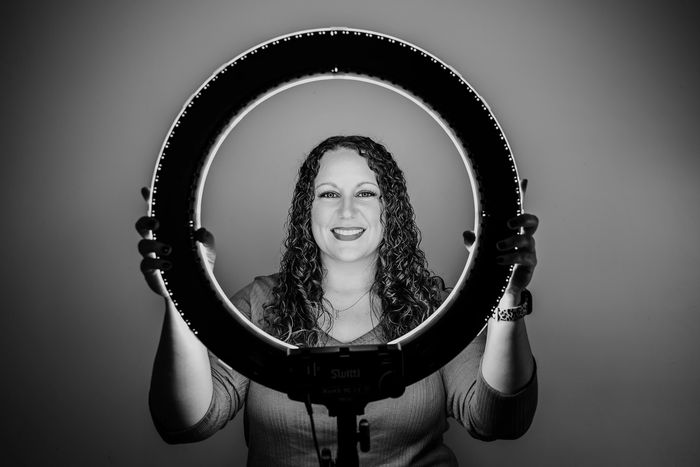 Woman smiling through a lit ring light in black and white.