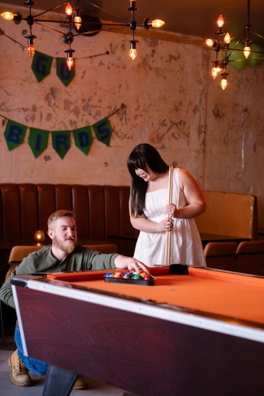 A couple setting up a pool game in a warmly lit room.