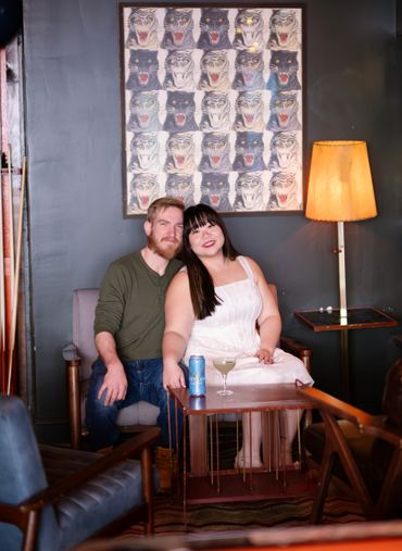 Couple sitting closely on a chair in a cozy room with a tiger artwork and warm lamp.