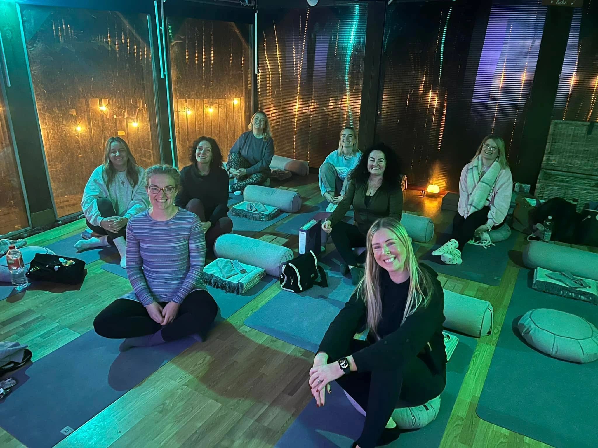 A group of women sitting on yoga mats in a dimly lit room with cozy lighting.