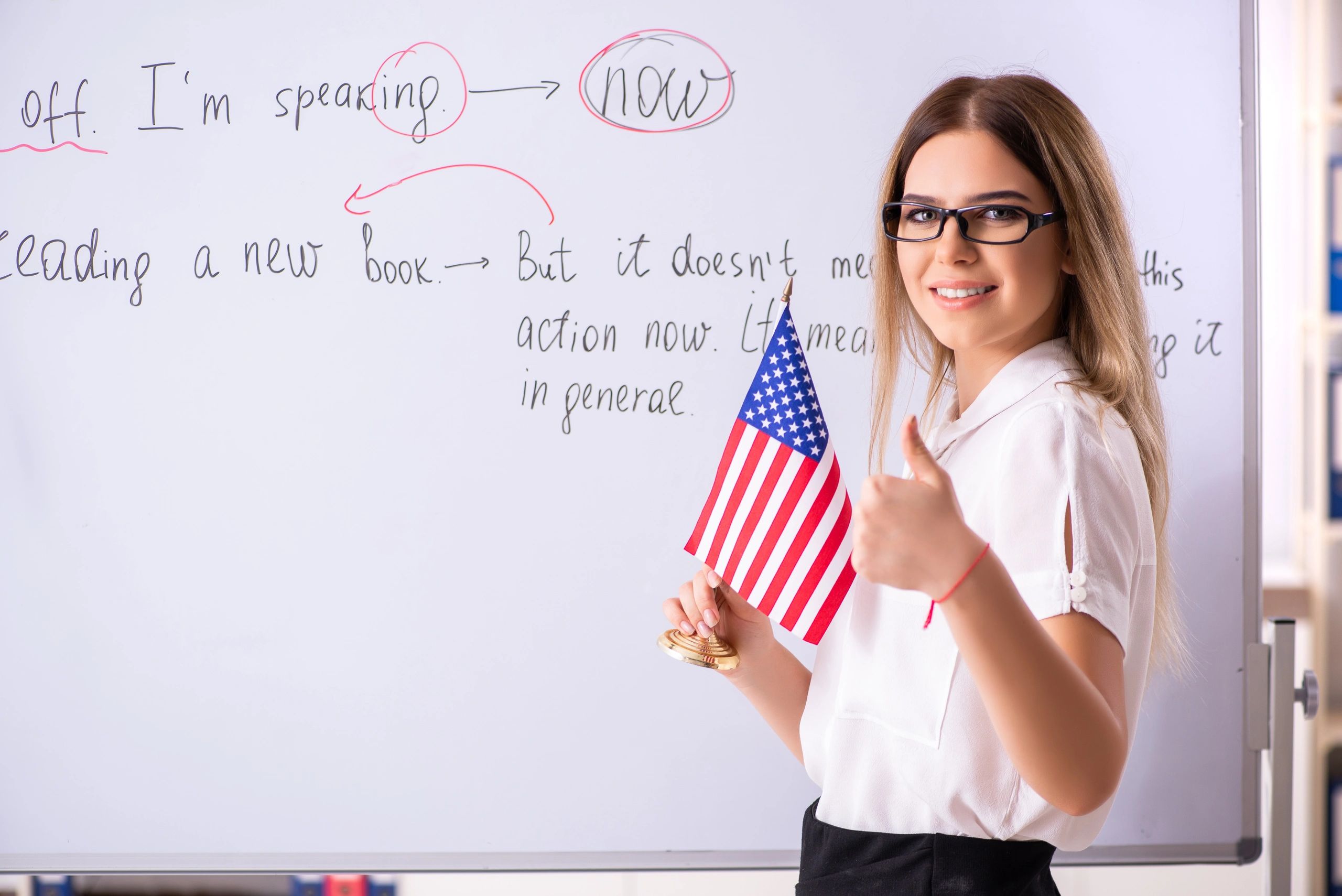 Teacher holding a U.S. flag gives a thumbs-up in front of a whiteboard with English lesson notes.