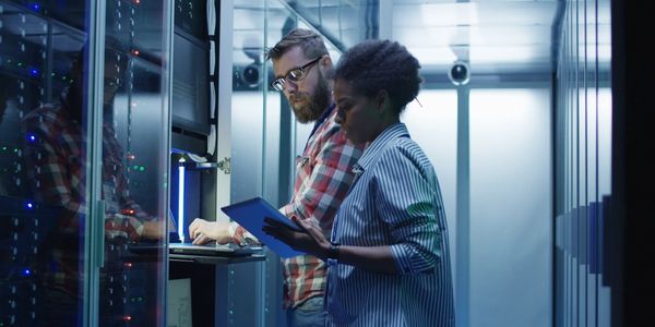 Two IT professionals working in a server room with a tablet and laptop.