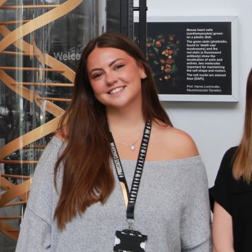 Smiling woman with long hair wearing a gray off-shoulder sweater and event lanyard.