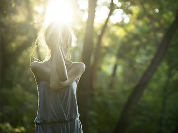 Woman standing in sunlight with hands behind her neck in a forest.