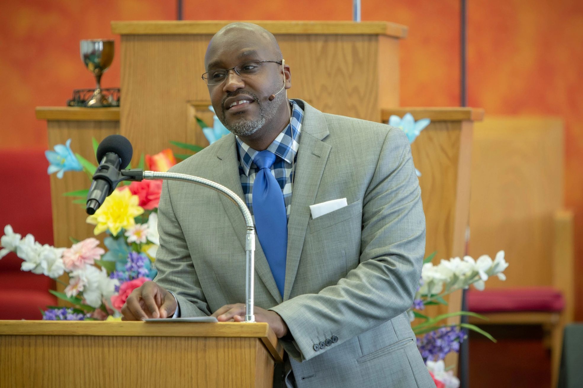 Man in suit speaking at a podium with flowers and a microphone.