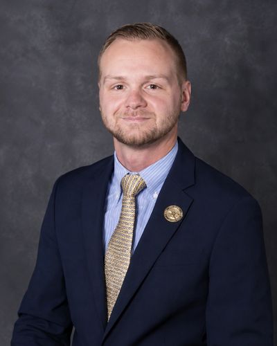 Professional portrait of a man in a navy suit and patterned tie against a gray backdrop.