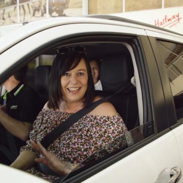 Smiling woman waving from inside a car wearing a floral top and seatbelt.