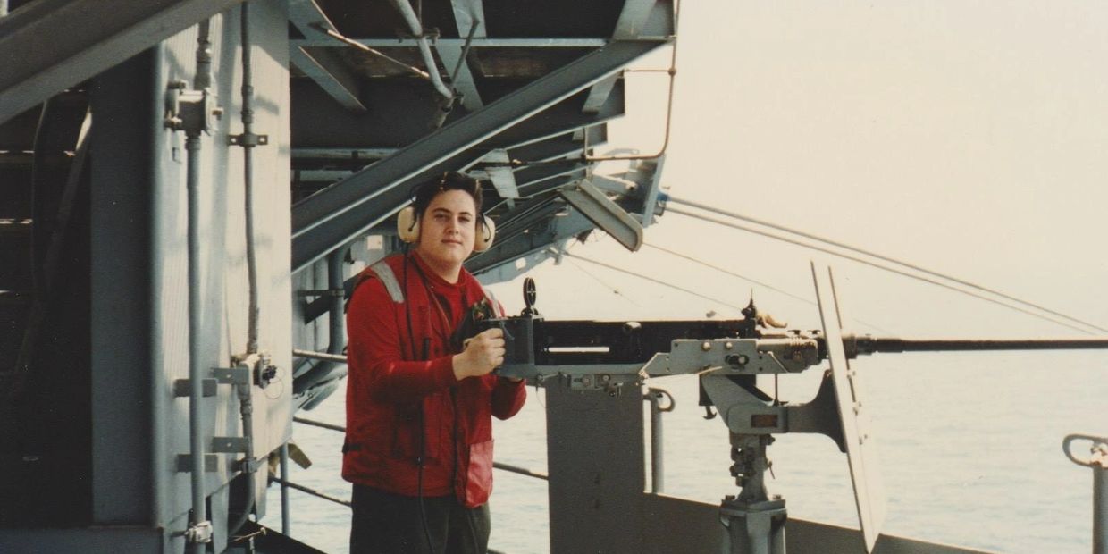 Person in red jacket operating a mounted machine gun on a ship deck.