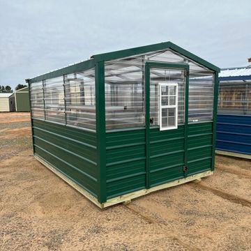 Green metal and glass greenhouse on a dirt surface under a cloudy sky.