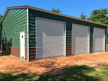 Green and brown metal garage with three white roll-up doors in a sunny yard.