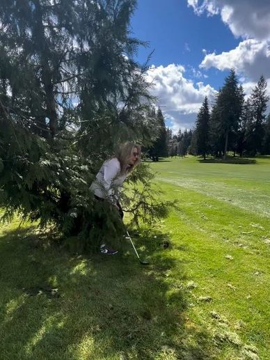 A woman playing golf hides behind a tree on a sunny day.