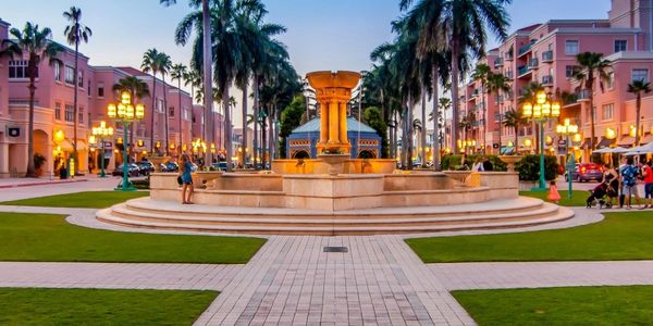 Evening view of a central fountain surrounded by palm trees and pink buildings with warm street lights.