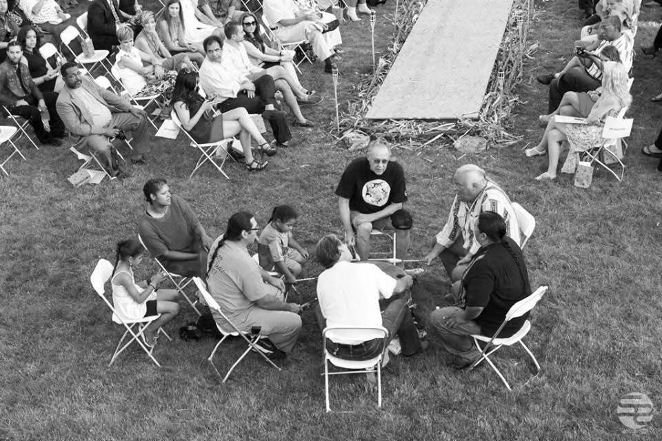 A group of people sitting in a circle on grass, surrounded by an audience.